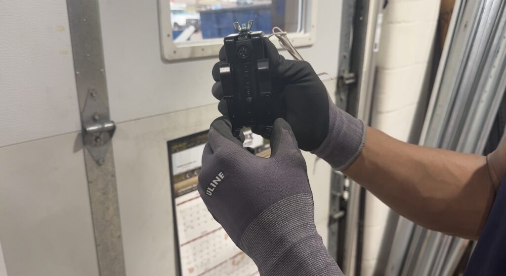 Technician wearing gloves holding and adjusting a garage door photo eye sensor inside a garage.