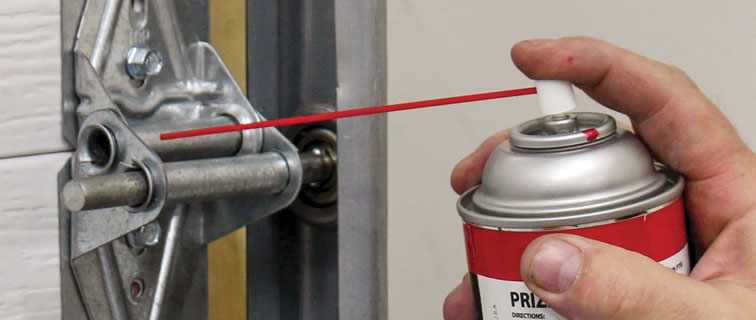 Hand spraying lubricant onto a garage door hinge and roller to reduce noise and improve performance.