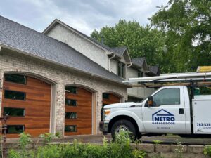Metro Garage Door service truck parked in front of a home with three modern wood-look garage doors featuring black windows after a completed installation.