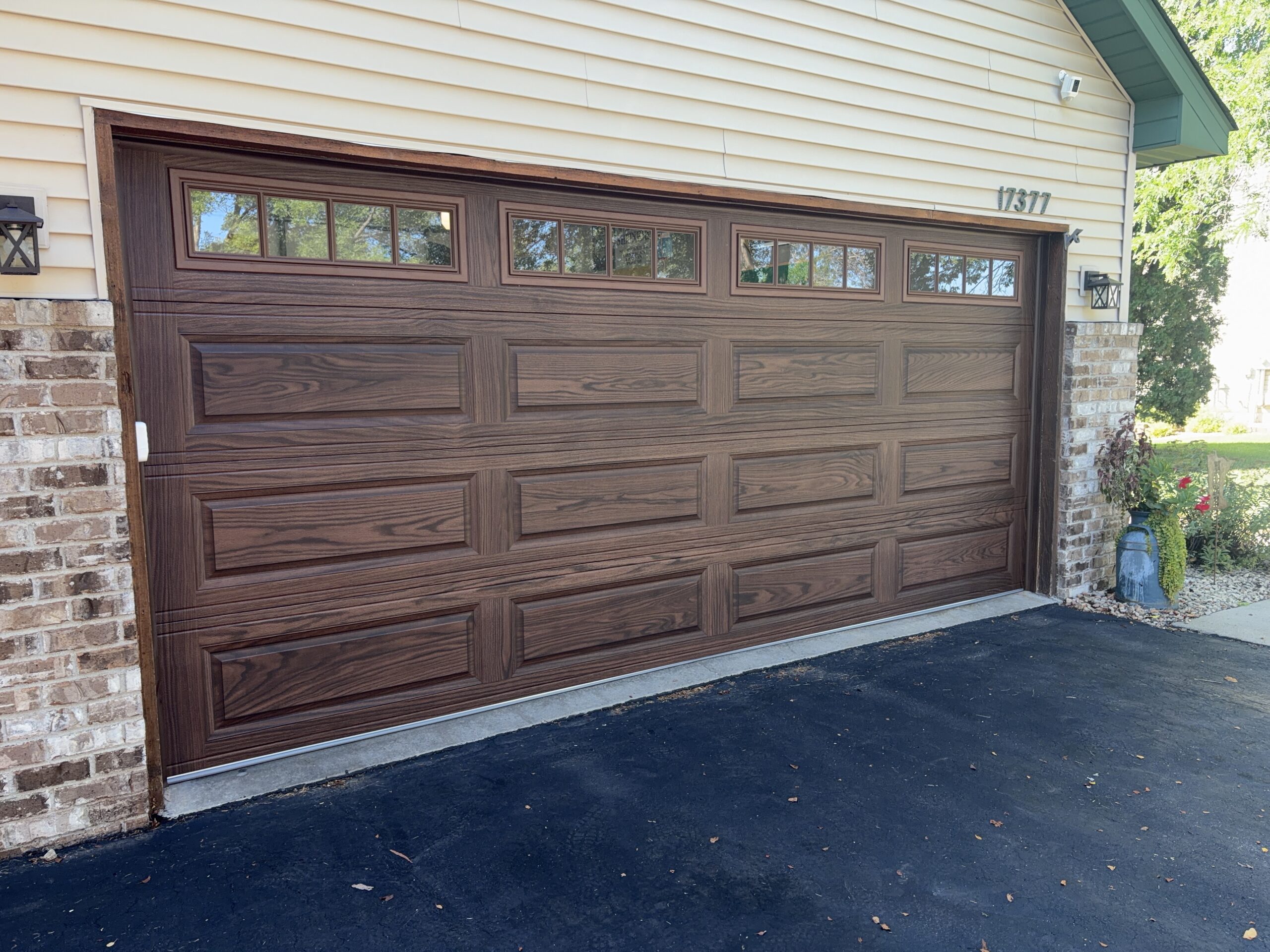 Brown wood-grain double garage door with raised rectangular panels and divided top-row windows, framed by brick and cream siding