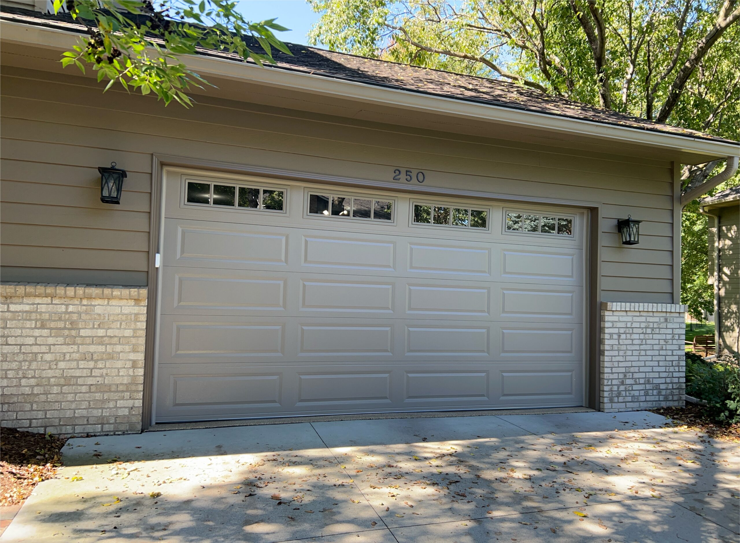 Beige double garage door with raised panels and divided top-row windows, surrounded by tan siding, light brick, and black lantern lights.