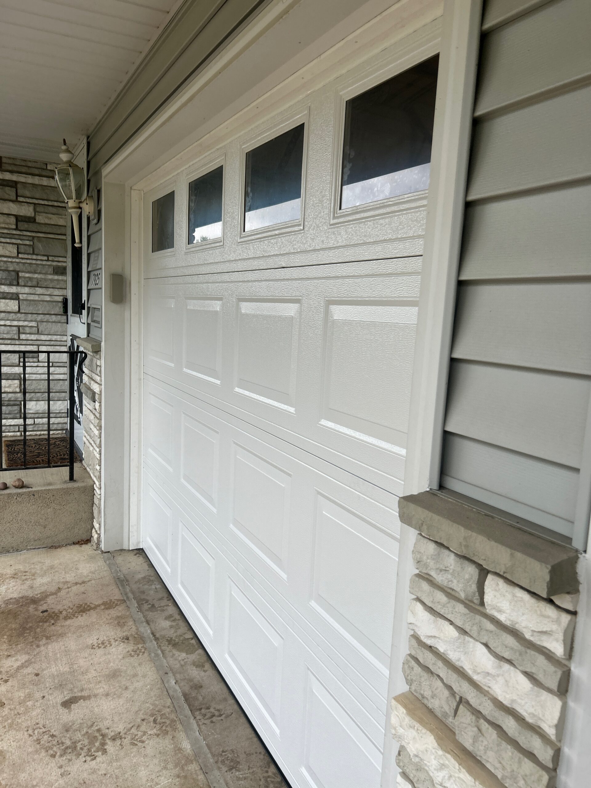 Beige double garage door with raised panels and divided top-row windows, surrounded by tan siding, light brick, and black lantern lights.