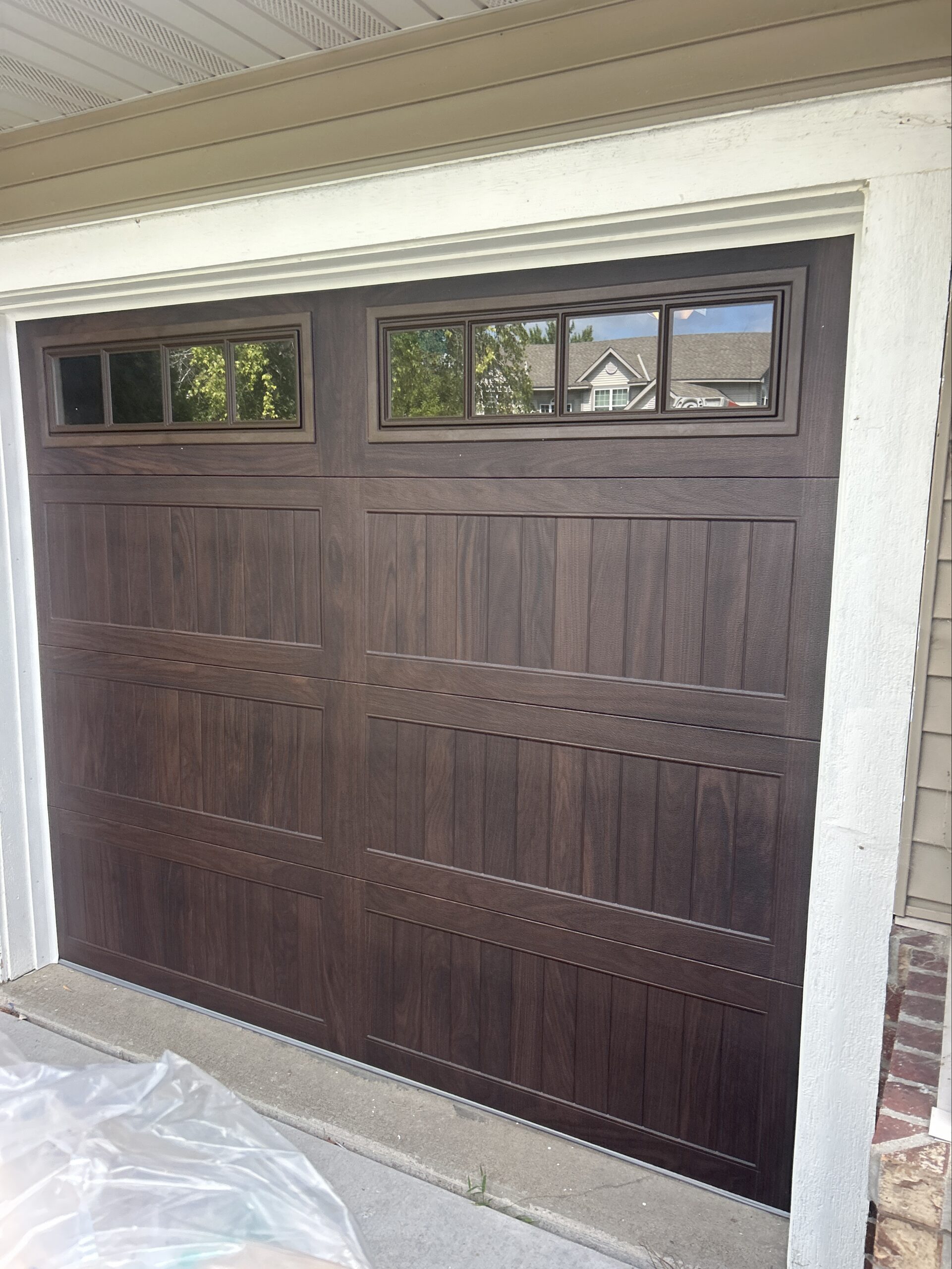 This double-width residential garage door features a textured brown finish, three rows of long raised rectangular panels, and four groups of divided windows across the top. Cream trim outlines the door against brown horizontal siding, with an asphalt driveway in front.