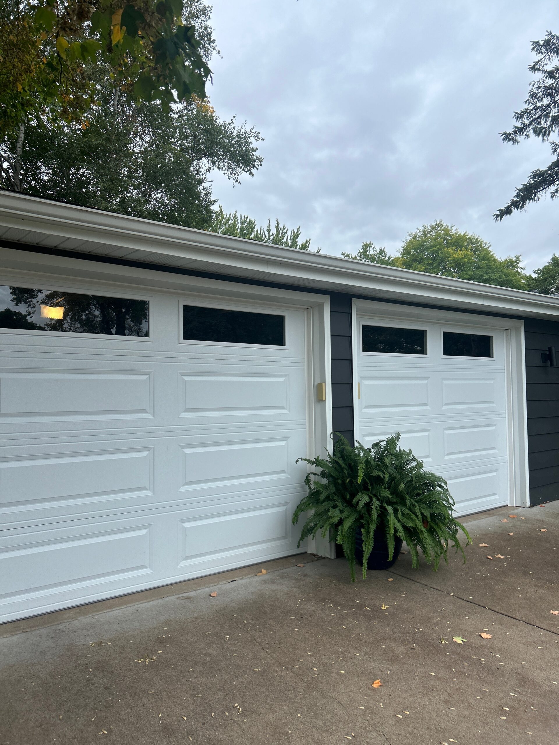 Two white single-stall garage doors with raised panels and rectangular top-row windows, set in dark gray siding with a potted fern between them.