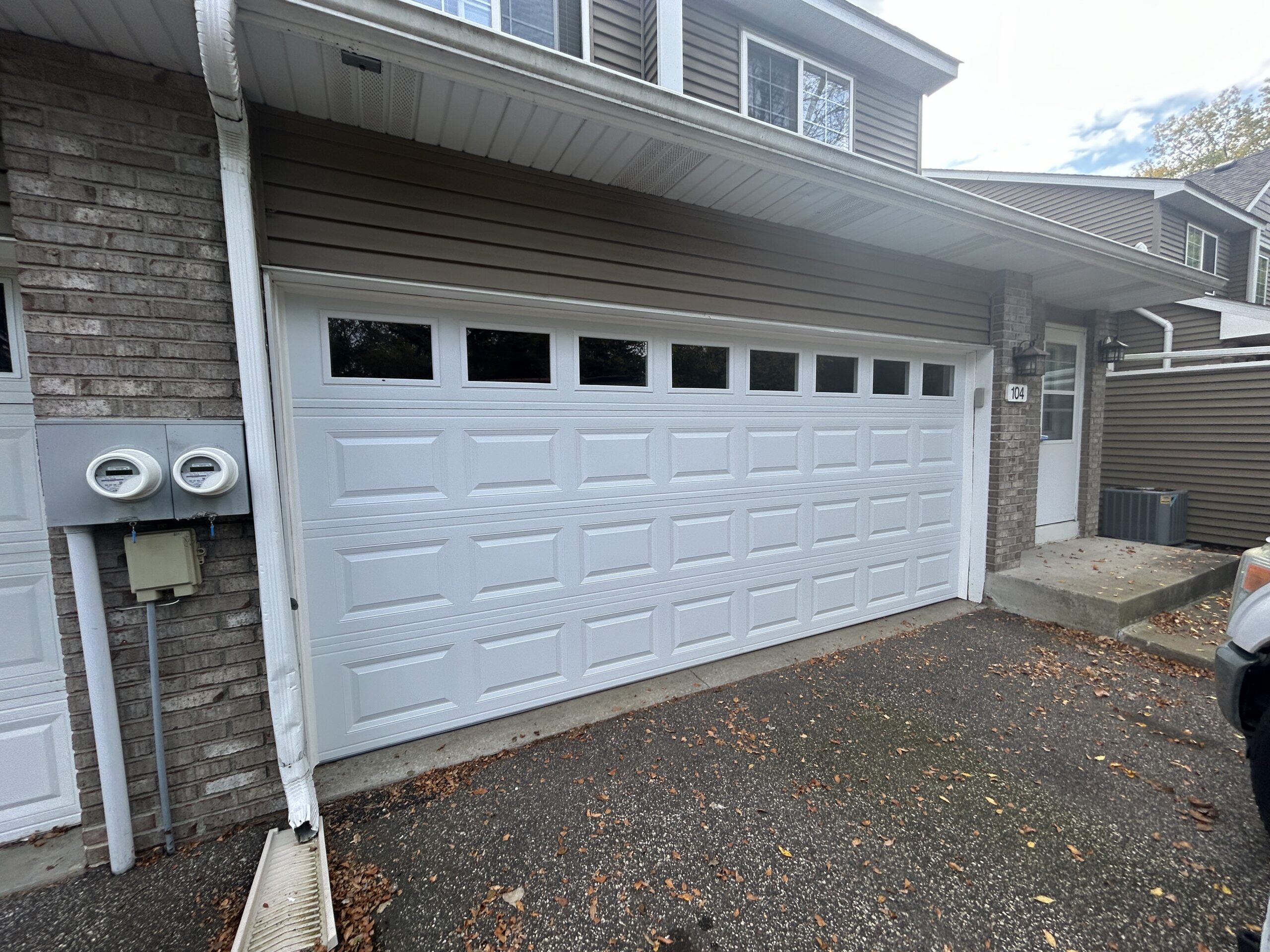 White double garage door with raised rectangular panels and eight top-row windows, framed by brick columns and brown siding.